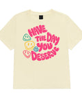 Buttermilk women's heavyweight garment-dyed tee, front view with pink Have the Day You Deserve lettering and colourful smiley faces.